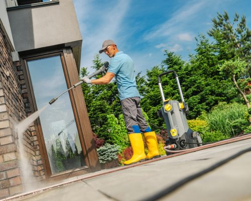 A man in yellow boots uses a pressure washer to clean the exterior walls of a home on a sunny day, surrounded by greenery and a clear blue sky.