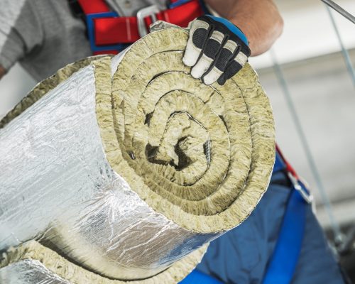 A construction worker holds rolled insulation material, preparing for installation. Safety gear is worn, highlighting the importance of worker safety on site.